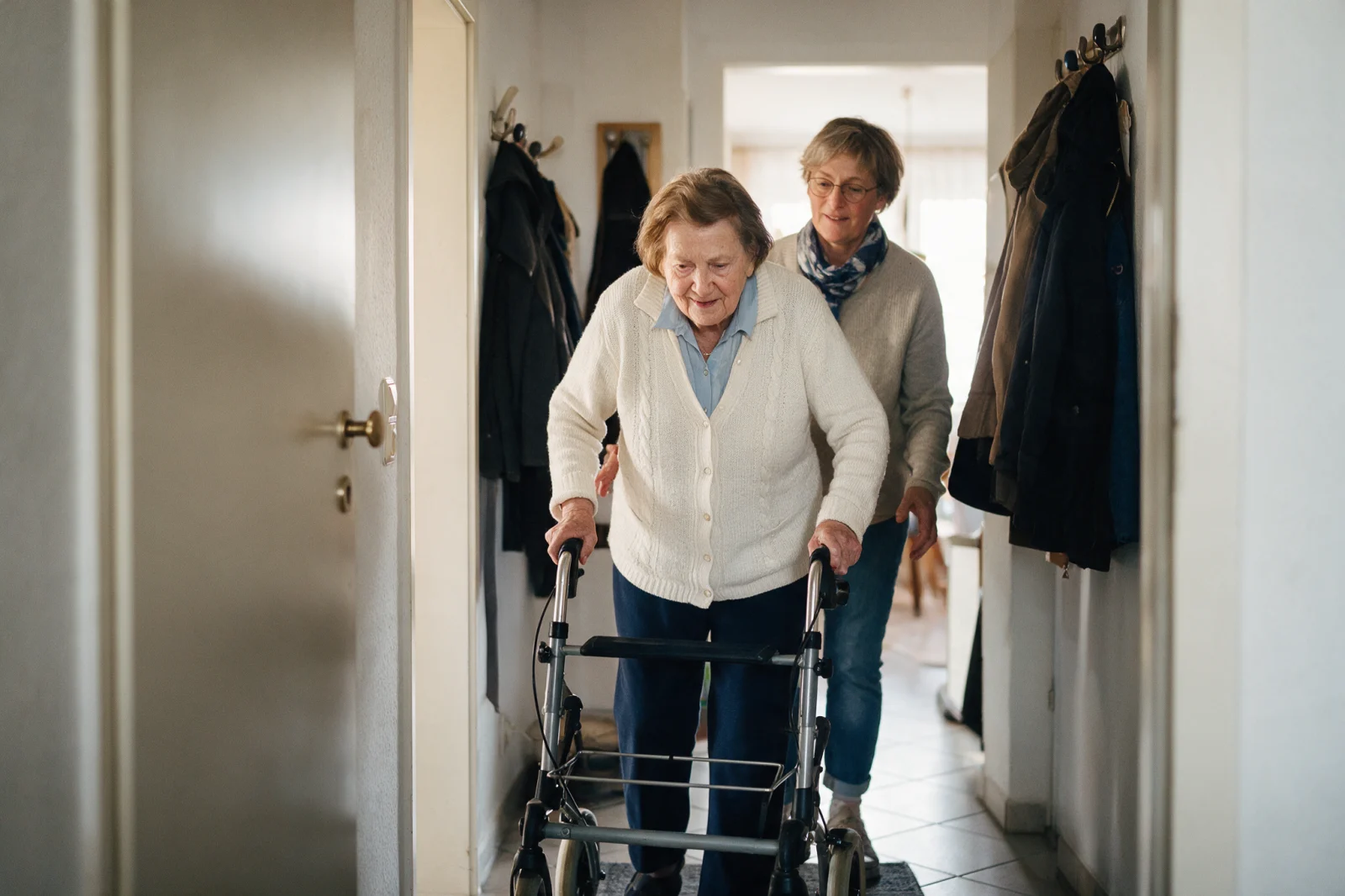 Ältere Frau geht mit Rollator durch ihren Flur, eine Angehörige begleitet sie